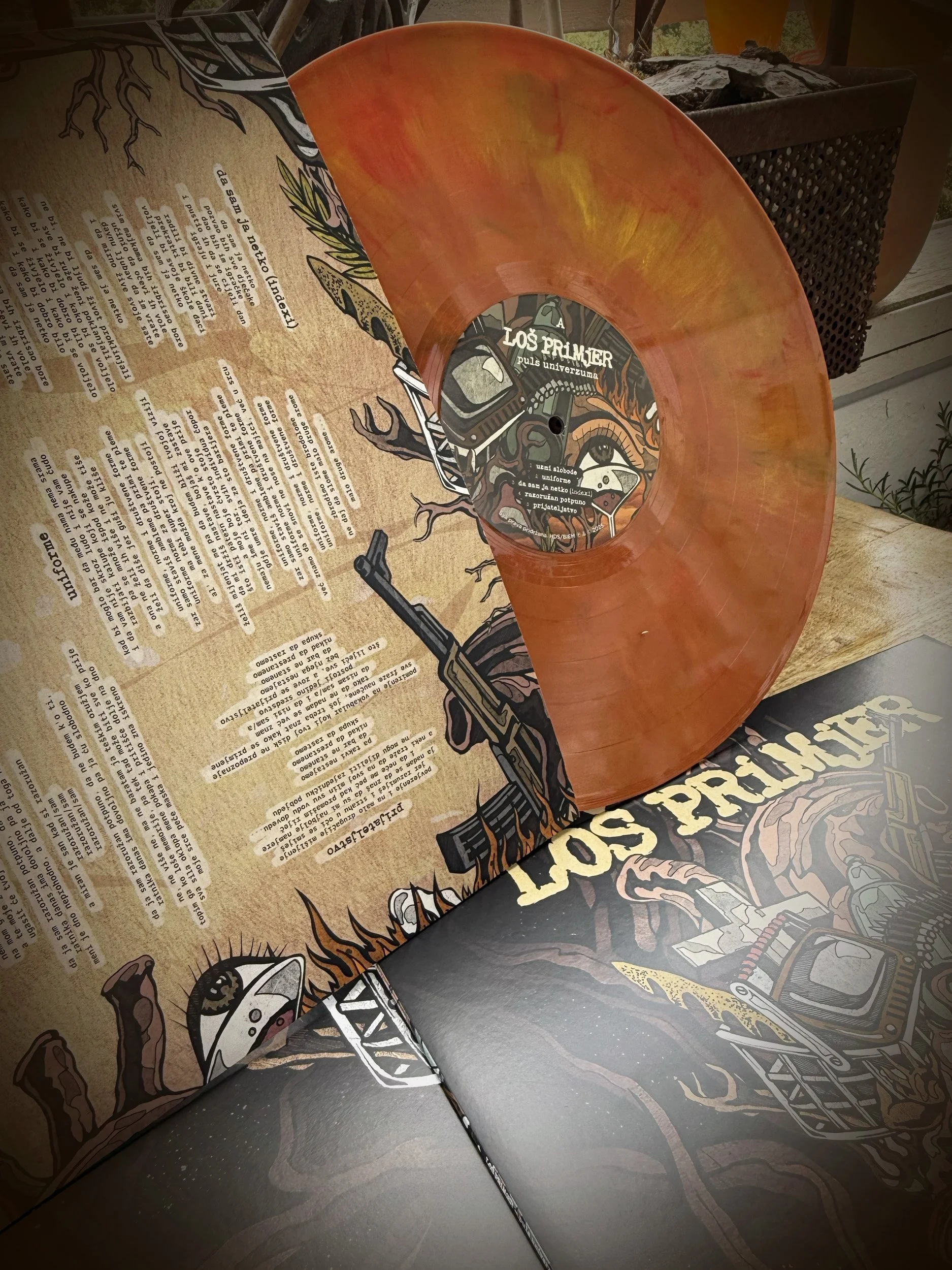 A vinyl record on top of a folded newspaper with illustrations and text, next to a grill with charcoal, and some potted plants.