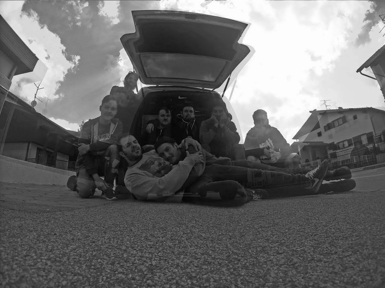 A group of nine people gathered around and sitting on the ground in front of an open car trunk, with some sitting inside the trunk, on a street in a residential neighborhood, with houses and cloudy sky visible in the background.