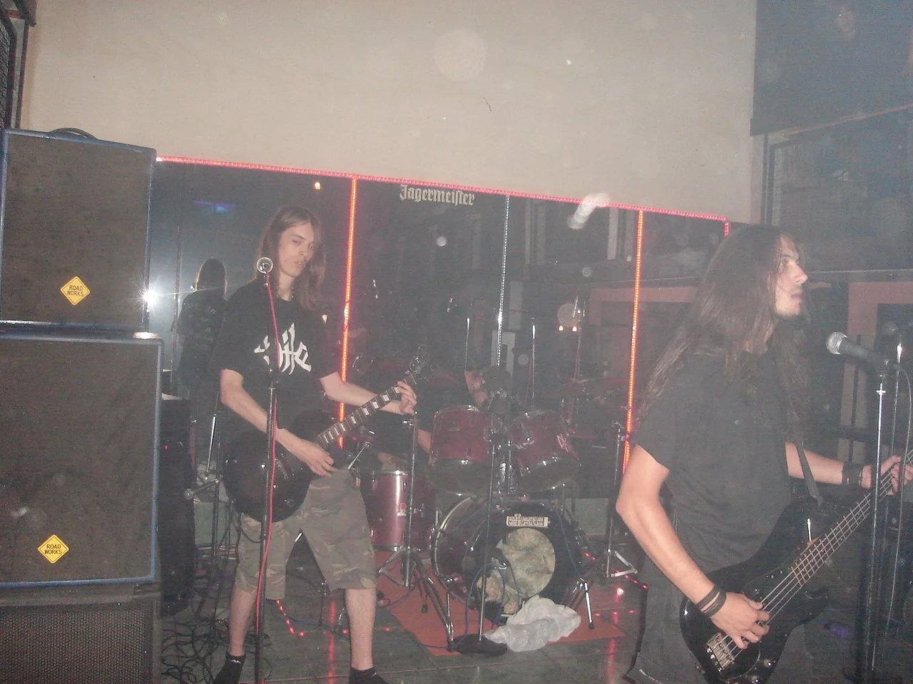 A band performing live on stage, featuring a female guitarist and bassist, with a drummer in the background, at a venue with a Jägermeister sign and neon lighting.