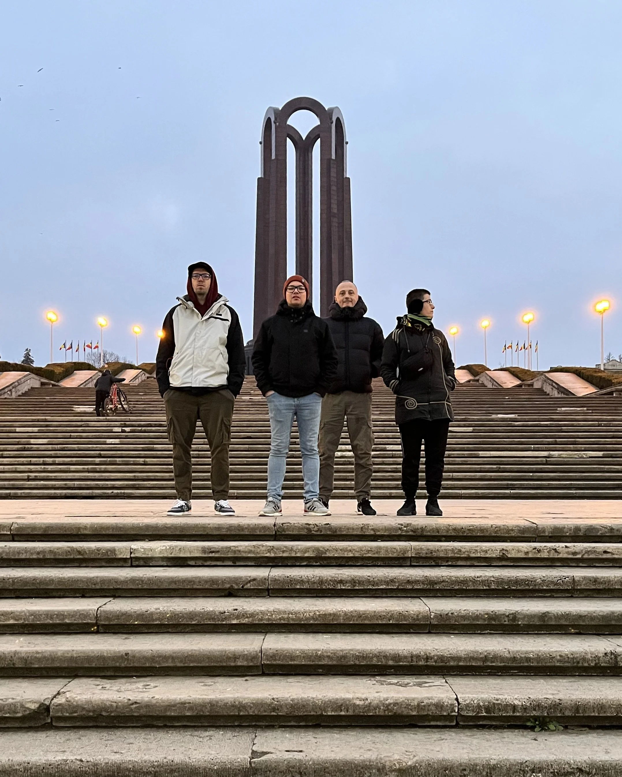 Four people standing on steps in front of a monument with skyscraper-like structure, surrounded by street lamps and flags, with a cloudy sky overhead.
