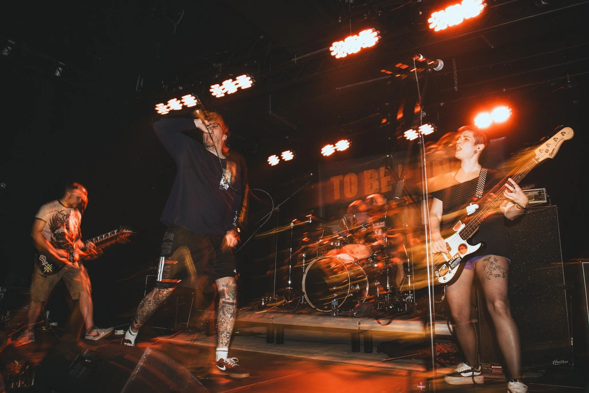 A live band performing on stage with lighting and colorful effects, featuring a male vocalist with glasses and tattooed legs, a female guitarist, and other band members in the background.