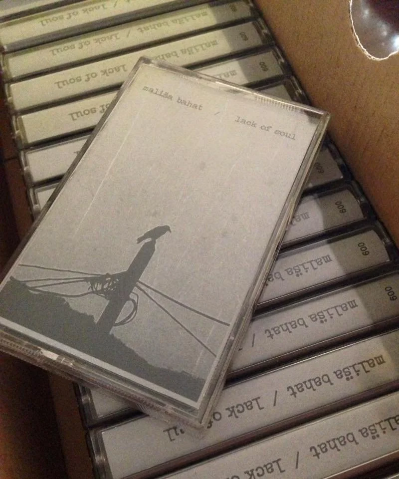 A cassette tape case with a cover featuring a silhouette of a bike and power lines, placed on a shelf among other cassette cases labeled 'Malisa Bane / Jack or Soul'.
