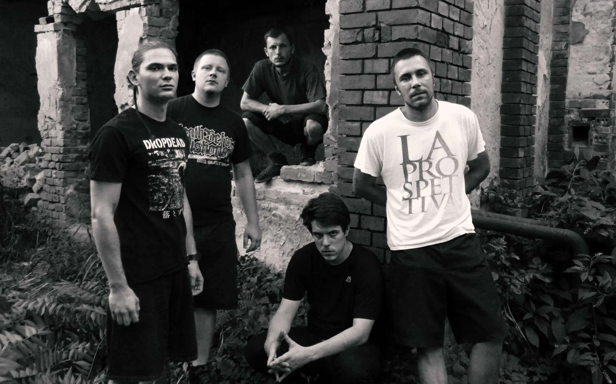 A group of six young men posing outdoors against a background of brick ruins and greenery. Some stand, two sit, and one crouches, with neutral or serious expressions.