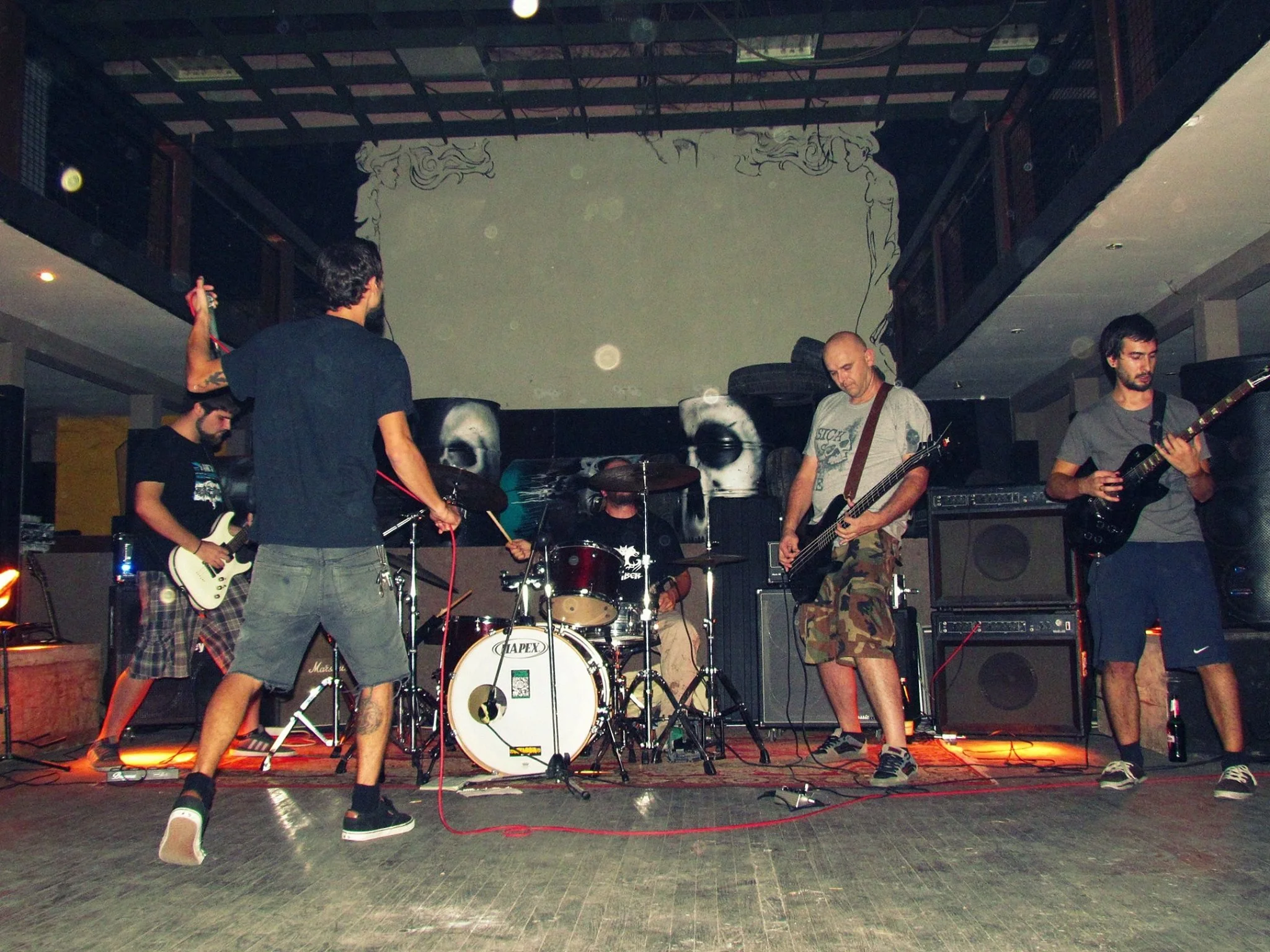 A band performing live on stage, with members playing guitars and drums, in a dimly lit venue with graffiti art and a skull image on the backdrop.