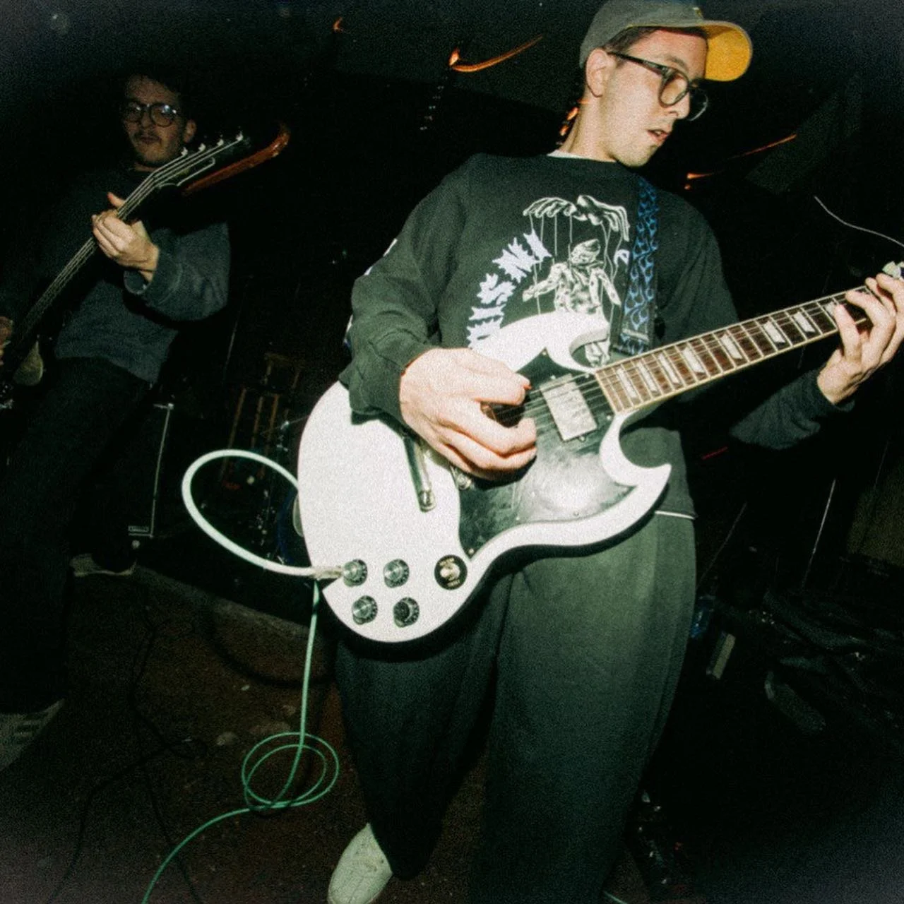 Two young men playing electric guitars on stage in a dimly lit venue, one wearing a baseball cap and glasses, the other in glasses and dark clothing.