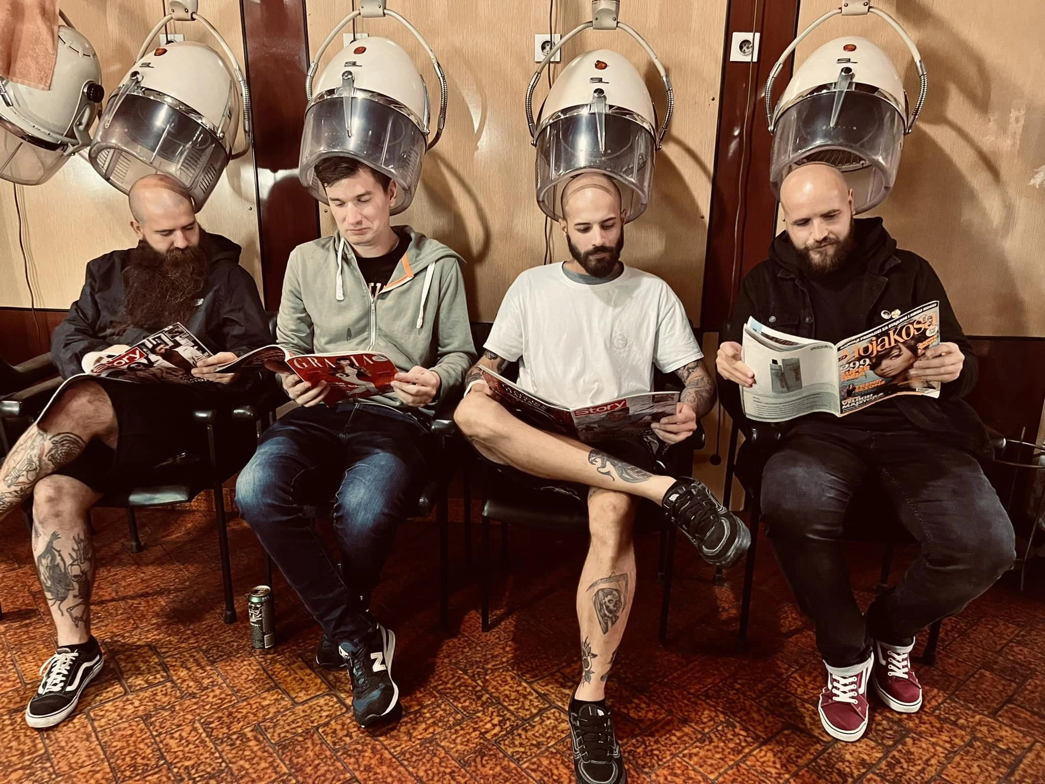 Four men with shaved heads and tattoos sitting in hair drying chairs with hood dryers over their heads, reading magazines in a salon or barbershop.