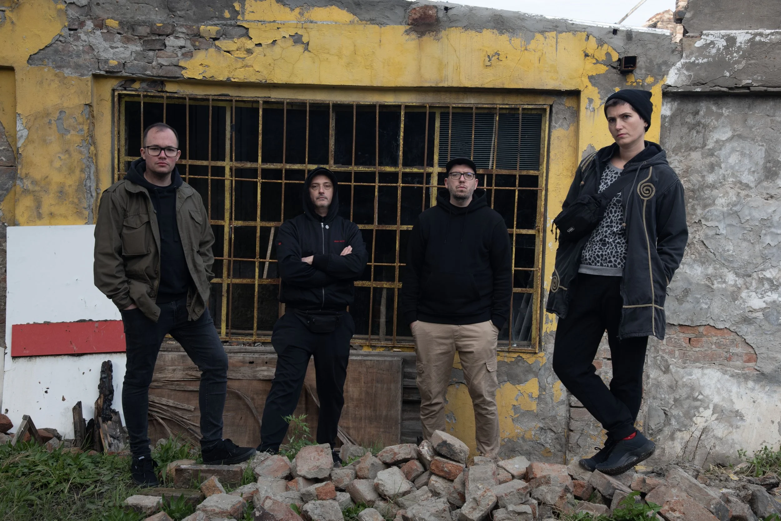 Four young adults standing in front of a dilapidated yellow wall with a barred window, surrounded by rubble and overgrown grass.