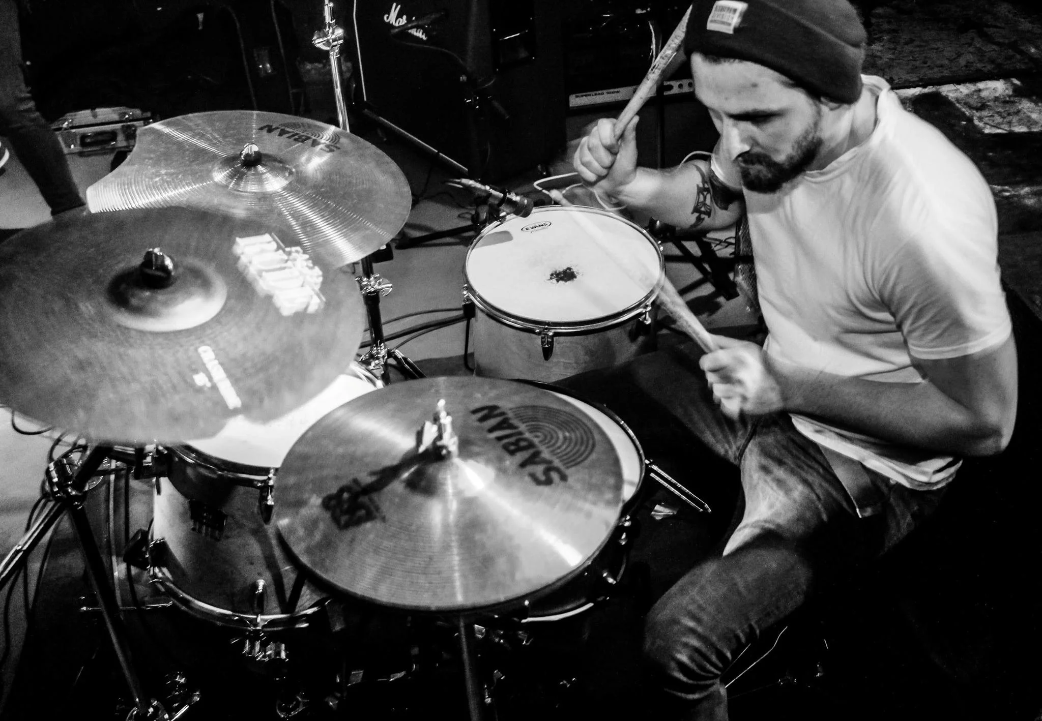 A man playing the drums, wearing a beanie and a white shirt, with a tattoo on his arm, surrounded by drum equipment.
