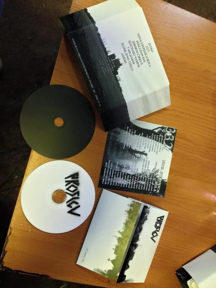 Two CDs, one black and one white with band logo, and two album covers, one with city skyline and the other with a group standing outdoors, arranged on a wooden table.