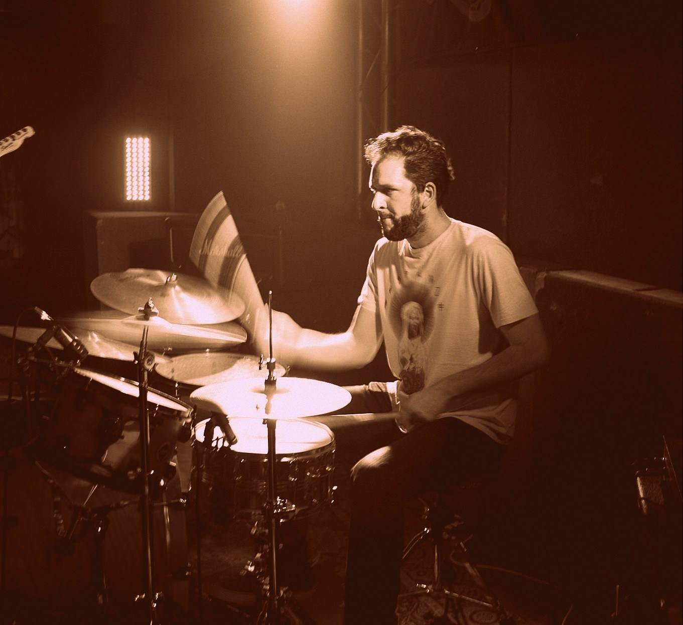 A man playing drums in a dimly lit music studio or stage, illuminated by warm lighting.