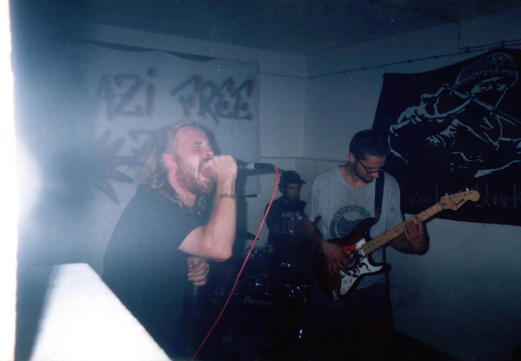 A band performs live in a dimly lit room; a man with long hair and beard sings into a microphone, and a guitarist plays an electric guitar in the background.