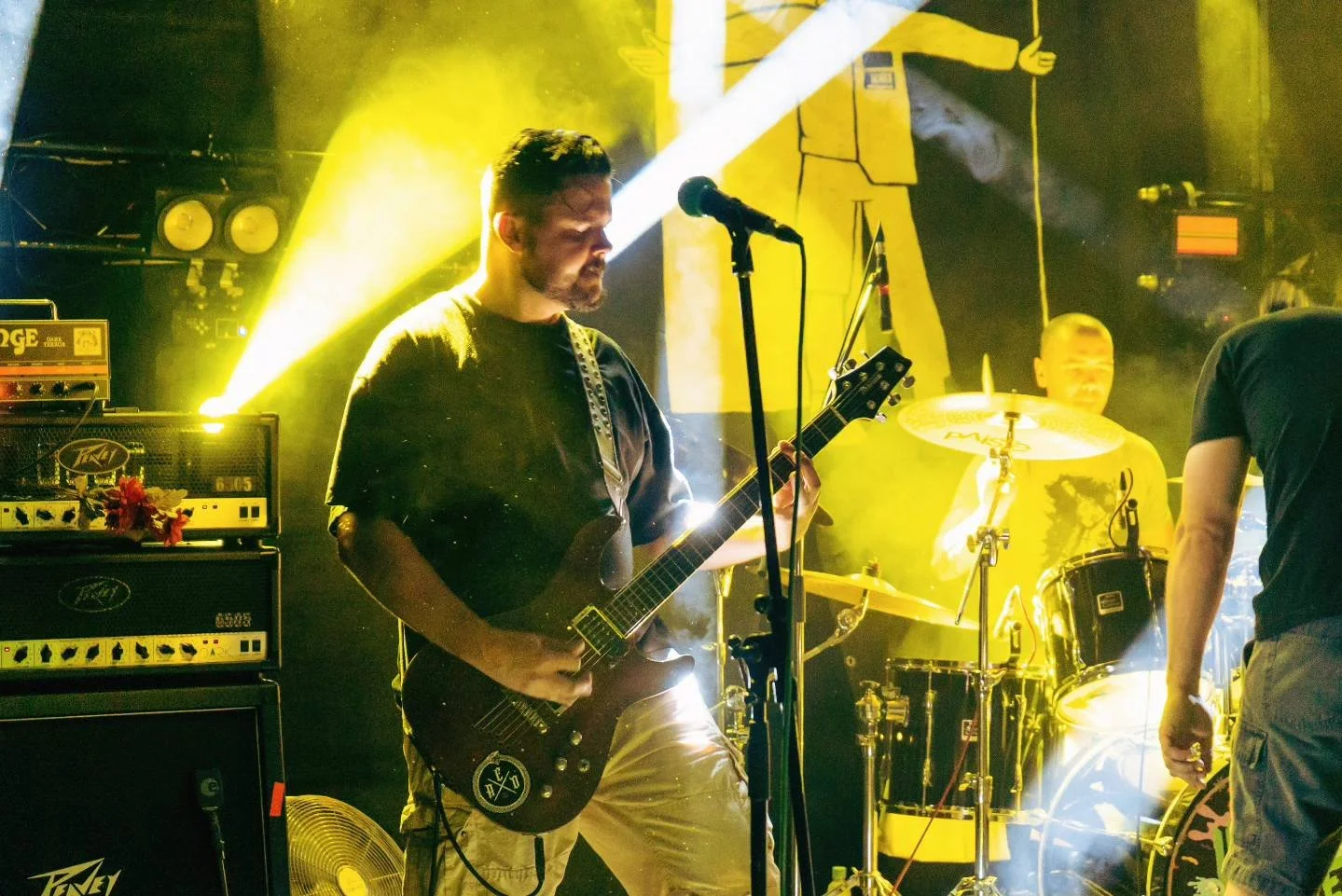 Musicians performing on stage with yellow lighting, one playing an electric guitar and another on drums, with equipment and stage lights in the background.