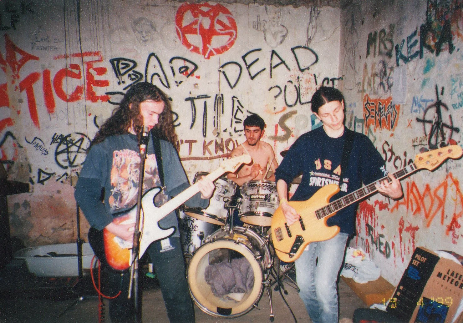 Three young musicians playing instruments in a room with graffiti-covered walls. One is playing an electric guitar, another on drums, and the third on bass guitar.
