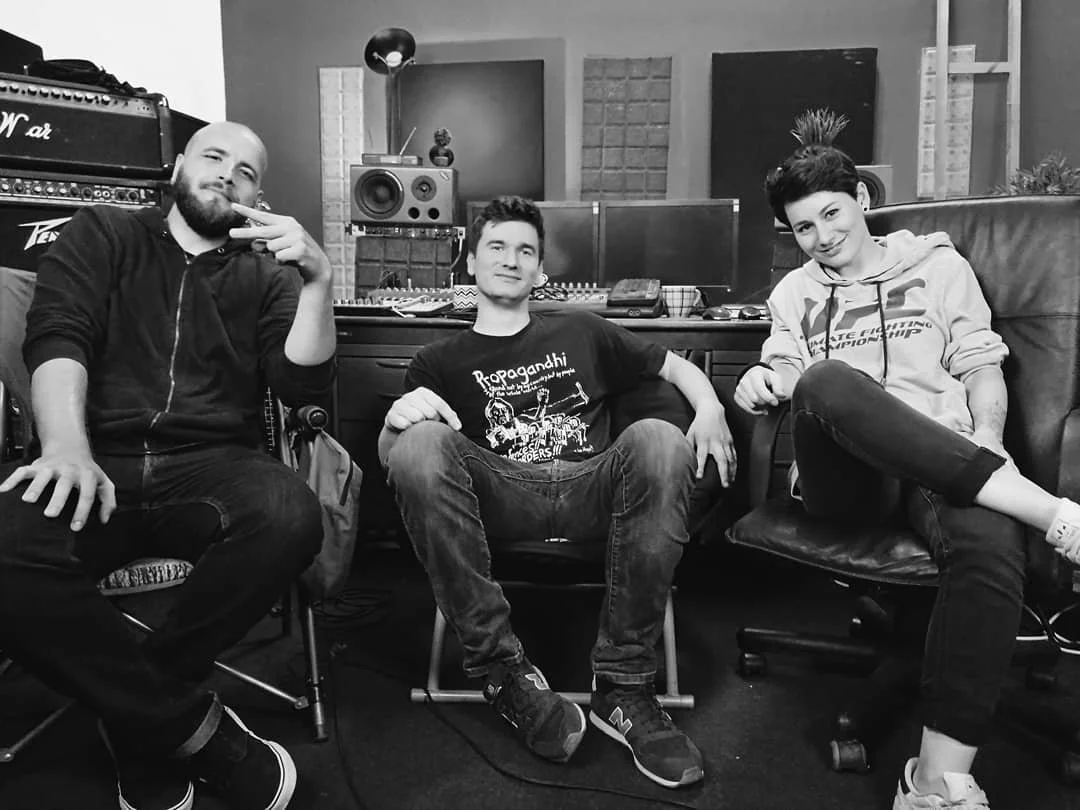 Three people sitting in a music studio surrounded by audio equipment and speakers, with one person making a peace sign.