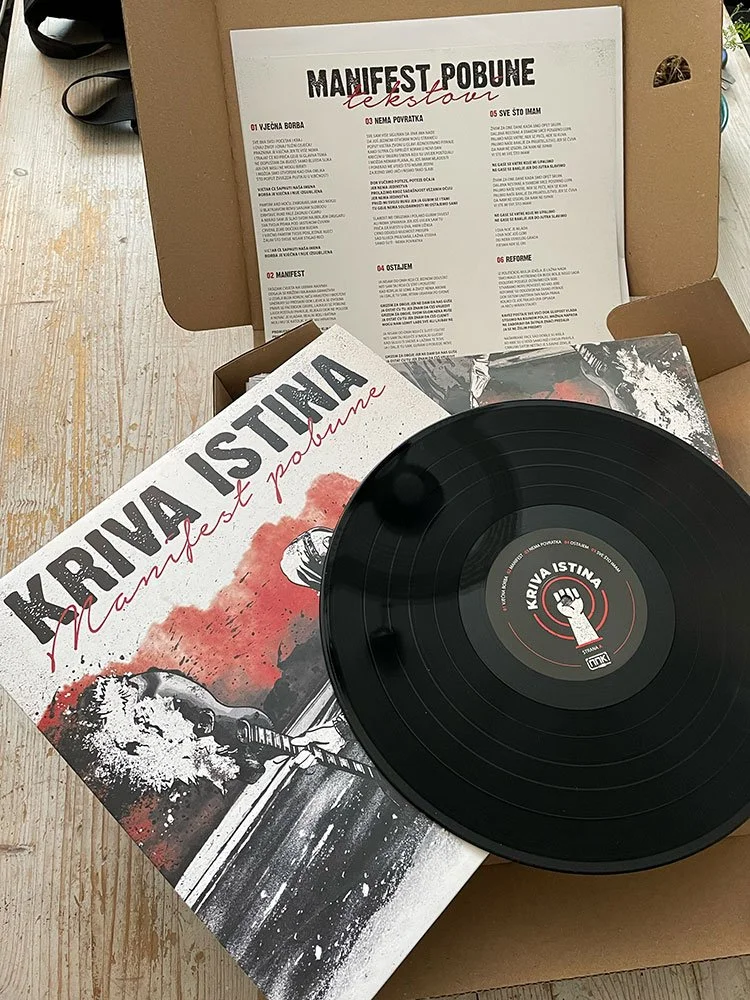 A vinyl record titled 'Kriva Istina Manifest Pobune' featuring artwork of a chain and a person's face. The record is inside a box along with an open pamphlet or manifesto titled 'Manifest Pobune' with six numbered sections and instructions.