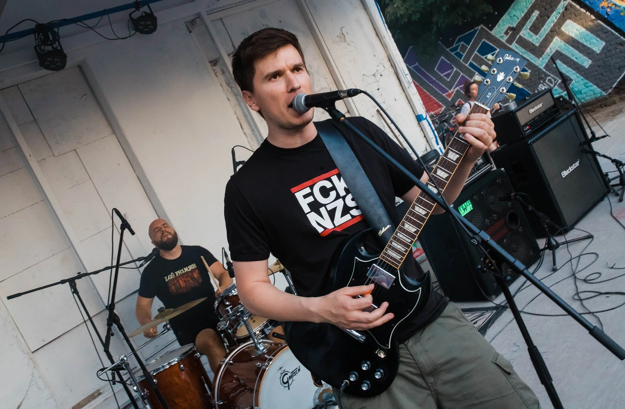 A band performing outdoors, with a singer playing an electric guitar and singing into a microphone, a drummer in the background, and a person playing bass guitar behind them. There are graffiti-covered walls behind the band.