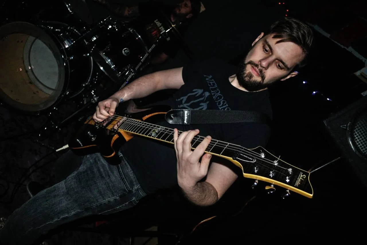 Young man playing electric guitar in a dark room with drums in the background.