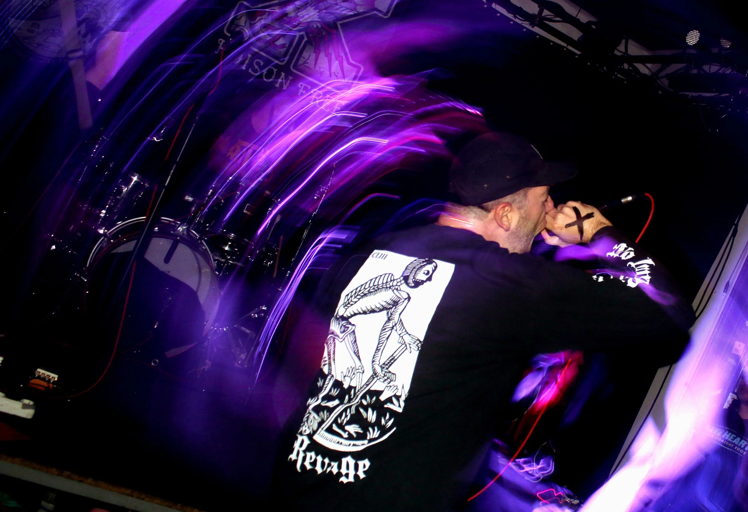 A person wearing a black hat and a black shirt with a white graphic design singing into a microphone on a stage with purple and pink light streaks.