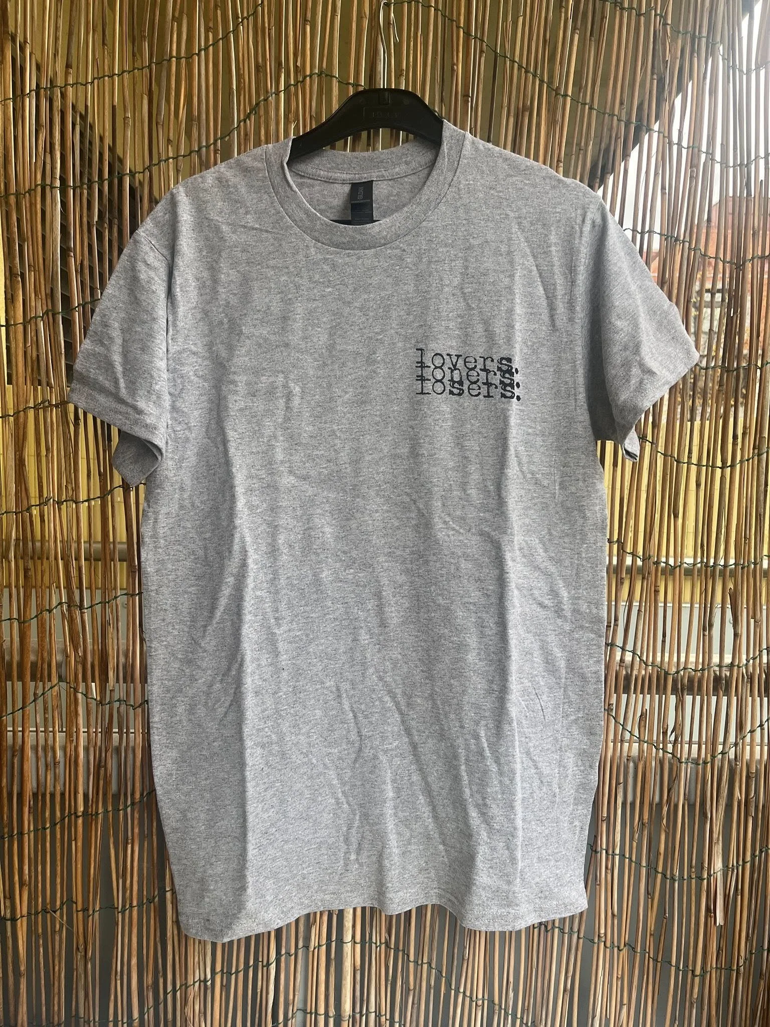 Gray T-shirt hanging on a black hanger against a bamboo background with the words "Lovers, Losers, loners" printed on the front.