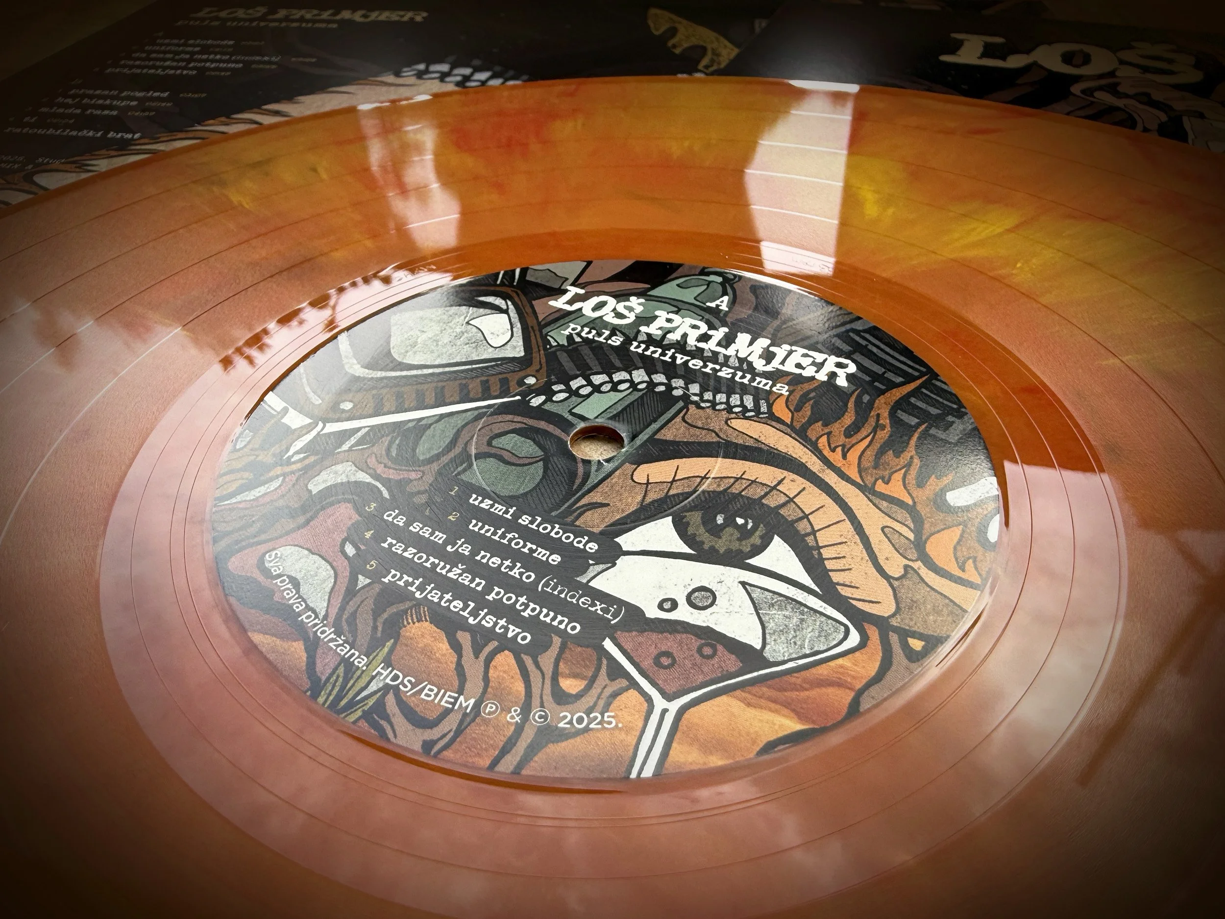 Close-up of a vinyl record with a colorful, illustrated label featuring a detailed, stylized eyeball and surrounding elements. The label has text in Polish and the record is partially resting on a black background with part of a magazine or booklet v