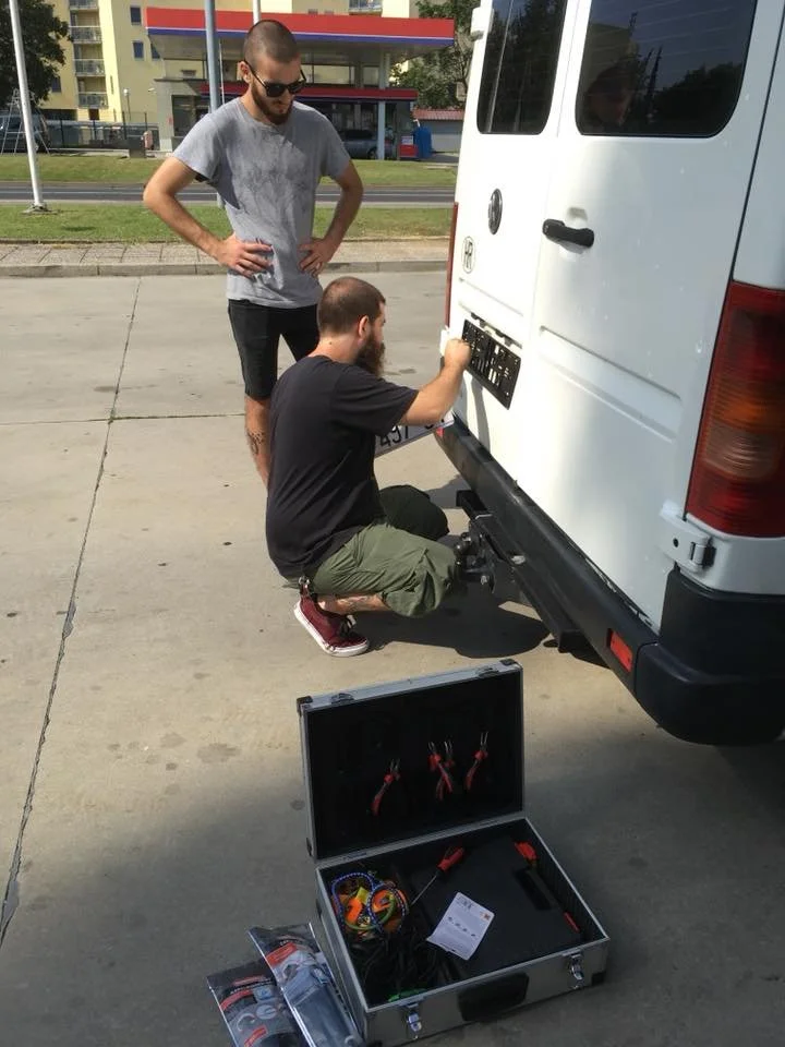 Two men work on installing a license plate on the back of a white van in a parking lot. One man is kneeling, using tools from an open toolbox, while the other stands nearby observing.