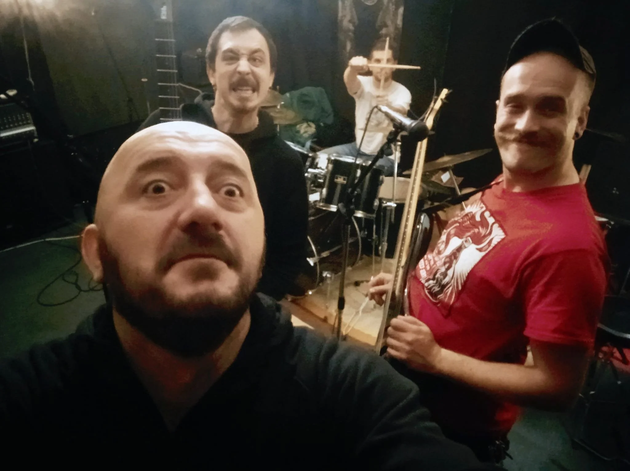 Four men in a music rehearsal room, with one person in the foreground taking a selfie. The man in the front has a bald head, a beard, and is making a surprised expression. The man on the right is smiling and holding a guitar. The man in the middle be