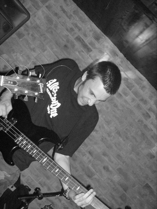 A man playing an electric guitar indoors, with a brick wall in the background.