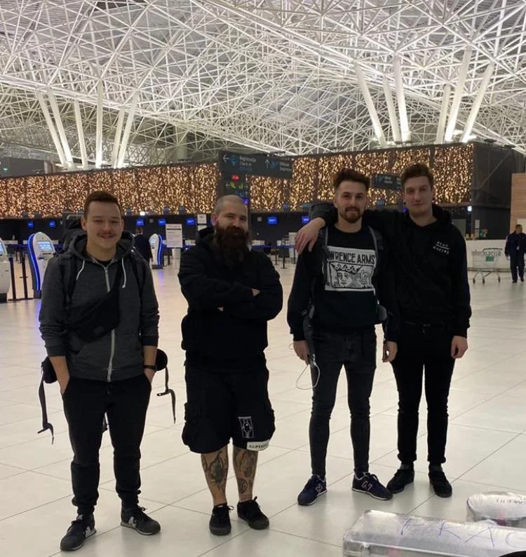 Four men standing inside an airport terminal with a metallic ceiling structure and a decorative gold-lit wall in the background.