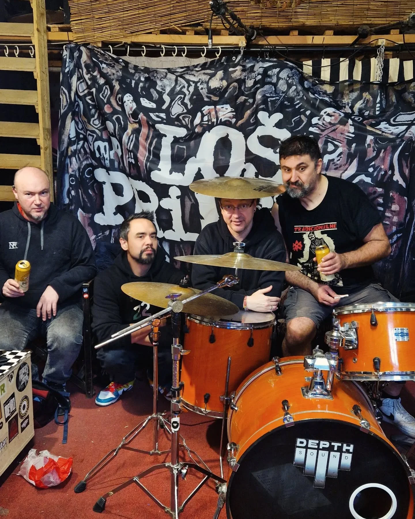 Four men sitting and posing around a bright orange drum set in a room with a black and white backdrop and wooden walls. Two of the men are holding cans of beverage, and one man is wearing a large sombrero hat. The backdrop has the words 'LOS P' print