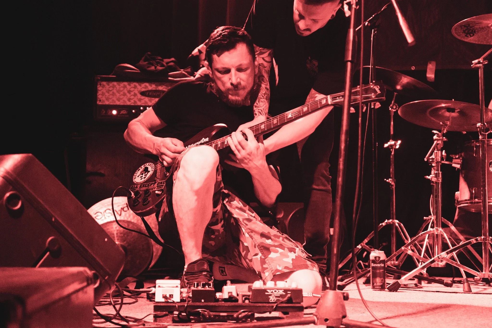 A musician playing an electric guitar on stage with another person bending over him. There are musical equipment and microphone stands around, with warm, red lighting. The musician is wearing camo shorts and a black T-shirt.