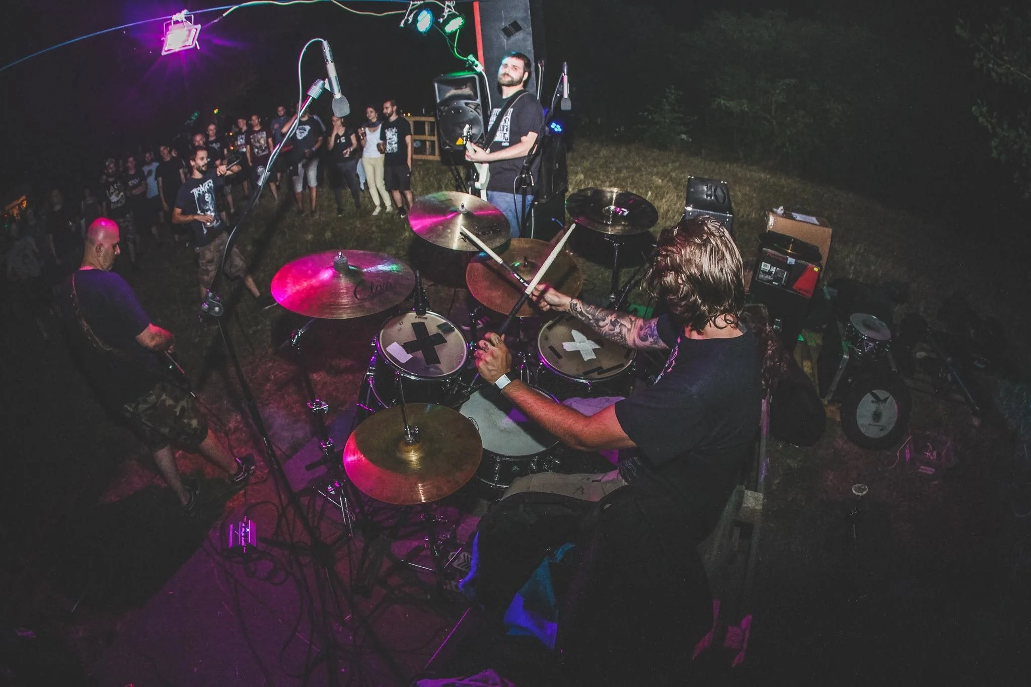 A live outdoor music performance at night with a drummer, a guitarist, and a guitarist in the background, surrounded by audience members. Colorful stage lights and equipment are visible.