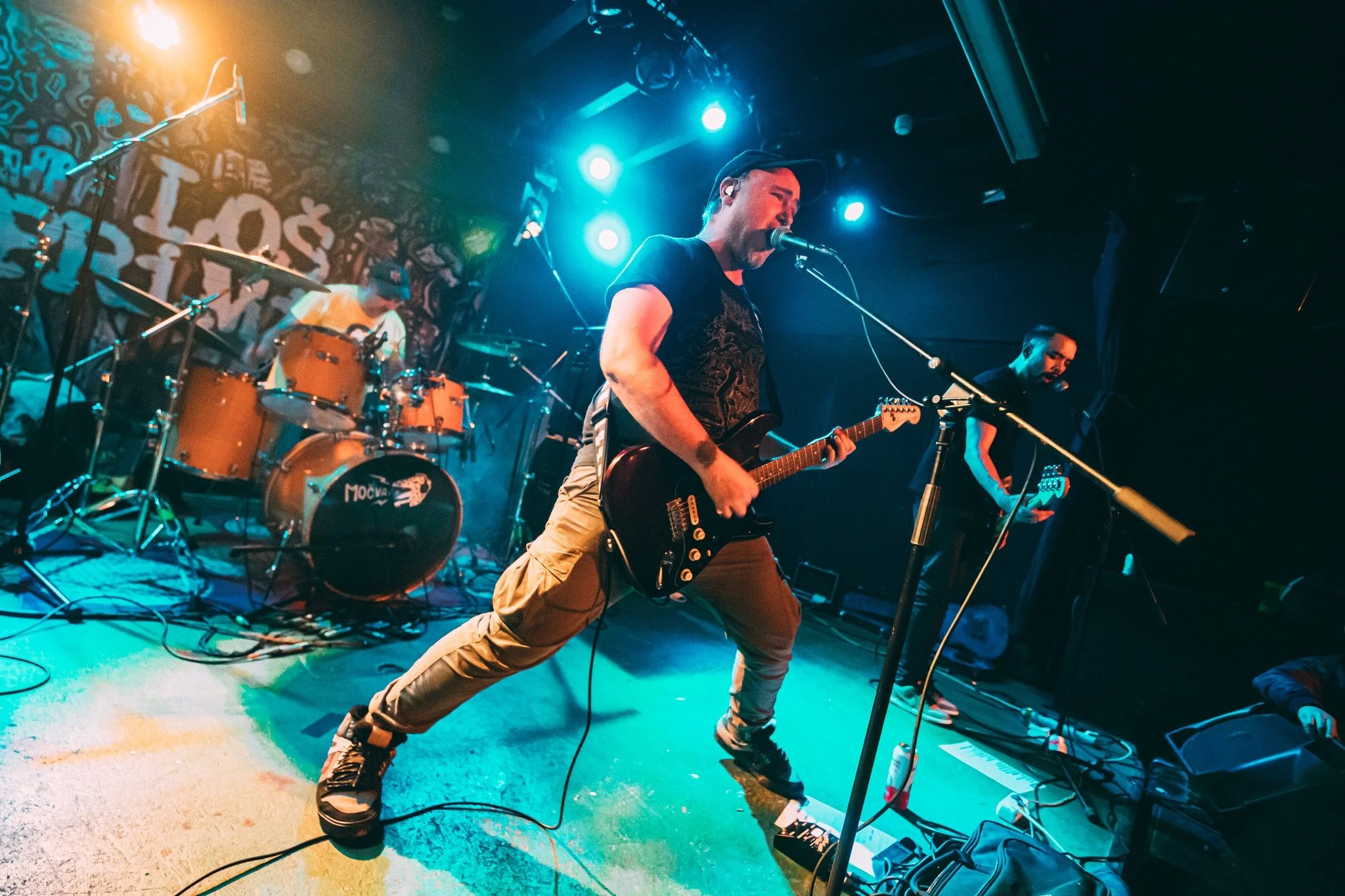 Musician playing electric guitar on stage during a live concert, with a drummer and another band member in the background, colorful stage lights, and a graffiti-style background.
