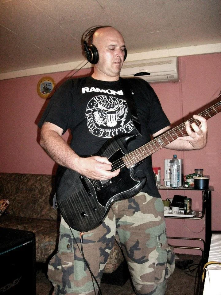 A man wearing a black Ramones T-shirt and camouflage cargo shorts is playing an electric guitar while listening to headphones in a room with pink walls and various items on a small table behind him.