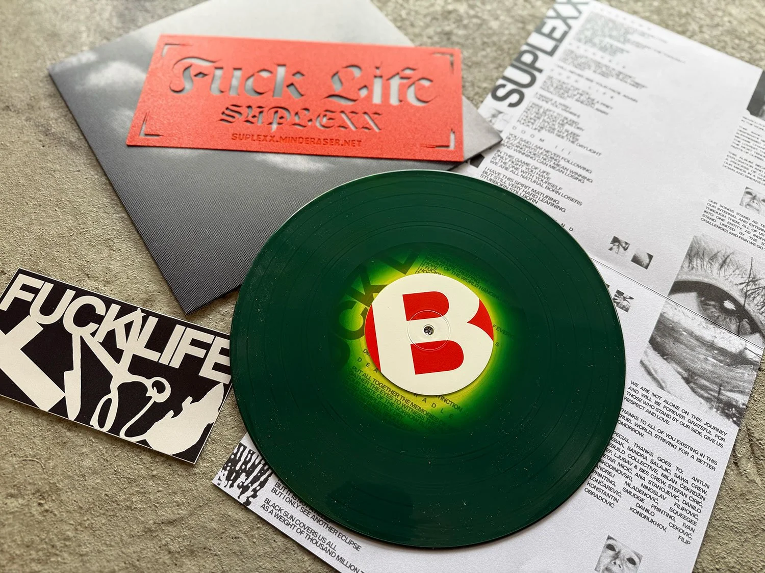 A green vinyl record with a large white and red letter B on the label, placed on top of a newspaper, accompanied by a gray envelope with a red cutout sticker that reads 'SUPLEXX', and a black and white sticker that says 'FUCK LIFE'.