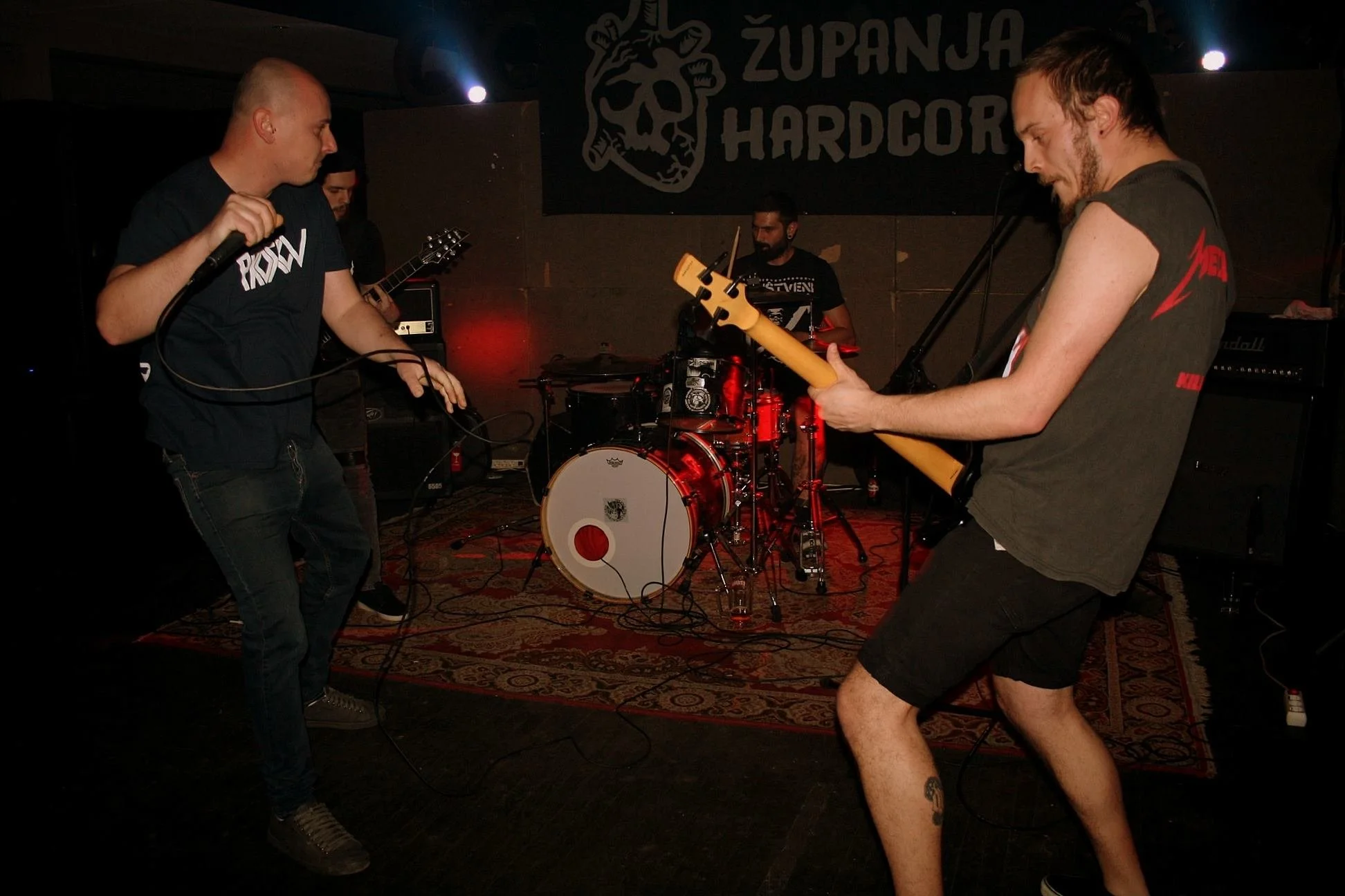 Four band members performing on stage at a venue with a banner saying 'Županja Hardcore' in the background. One member is holding a microphone, another is playing a guitar, the third is on drums, and the fourth is playing a bass guitar.