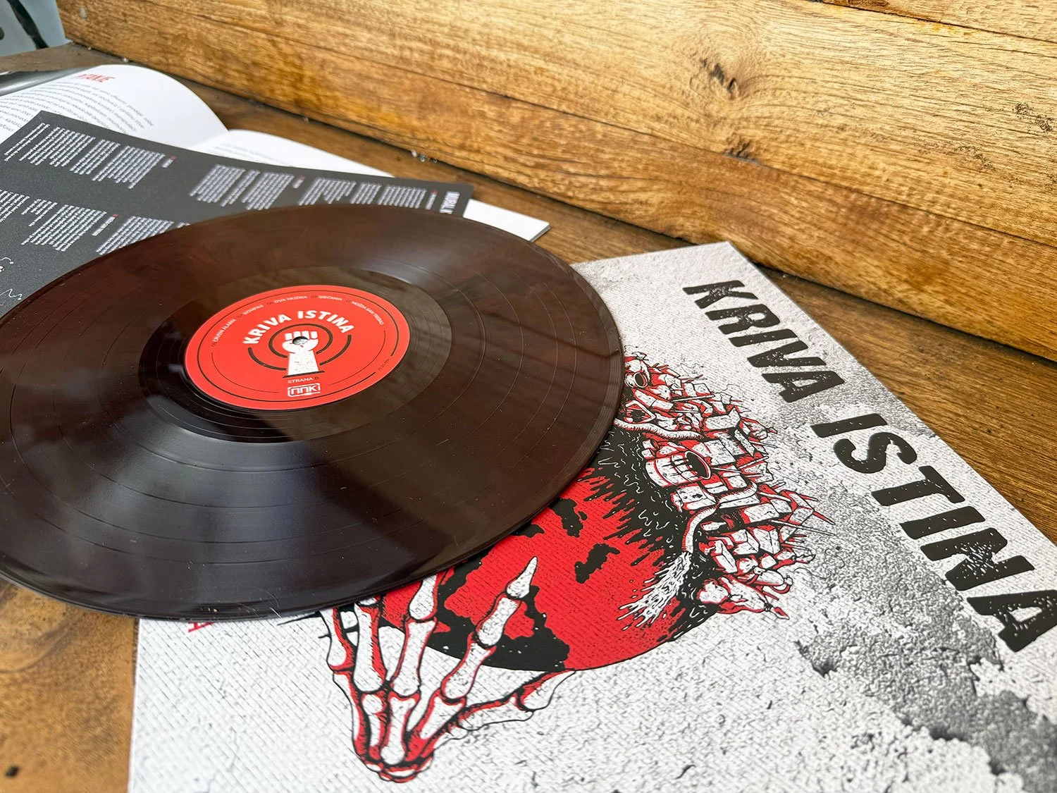 A black vinyl record with a red label resting on a cardboard cover with a cartoonish illustration of a zombie and the words "Krvava Istina" on it, placed on a wooden surface.