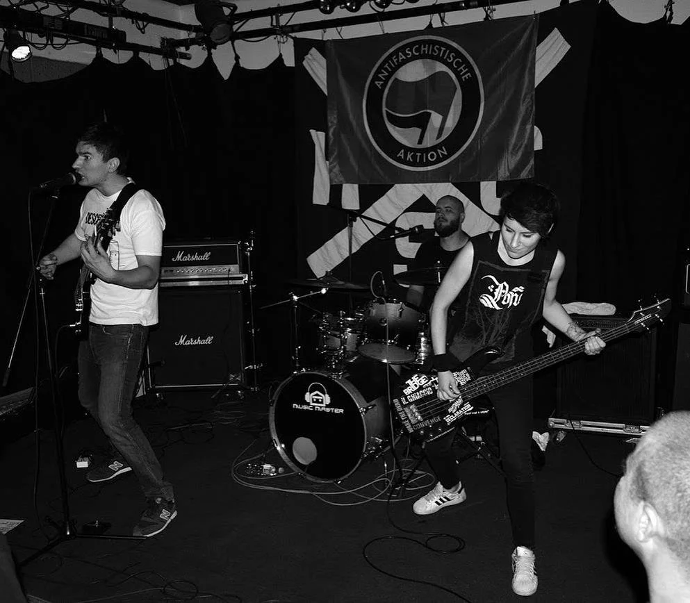 A band performing on stage with three members: a singer with a guitar, a drummer, and a bassist. The stage has a banner with the logo of the Antifascist Action in the background.