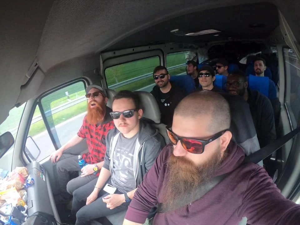 A group of eight men with varied appearances, some with beards and sunglasses, sitting inside a bus during a road trip. The bus is moving, with a highway and green scenery visible outside the windows.