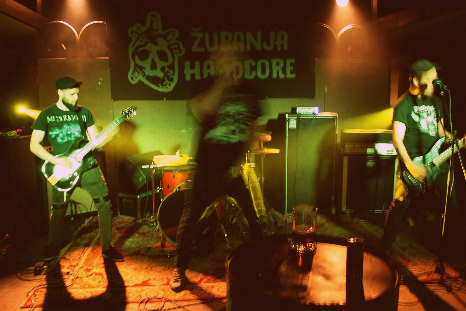 A band performing on stage at a live music venue with a banner behind them that reads 'ŽUPANJA HARDCORE' featuring a skull logo. The stage is lit with yellow and orange lights, and three band members are visible, playing guitars and drums.