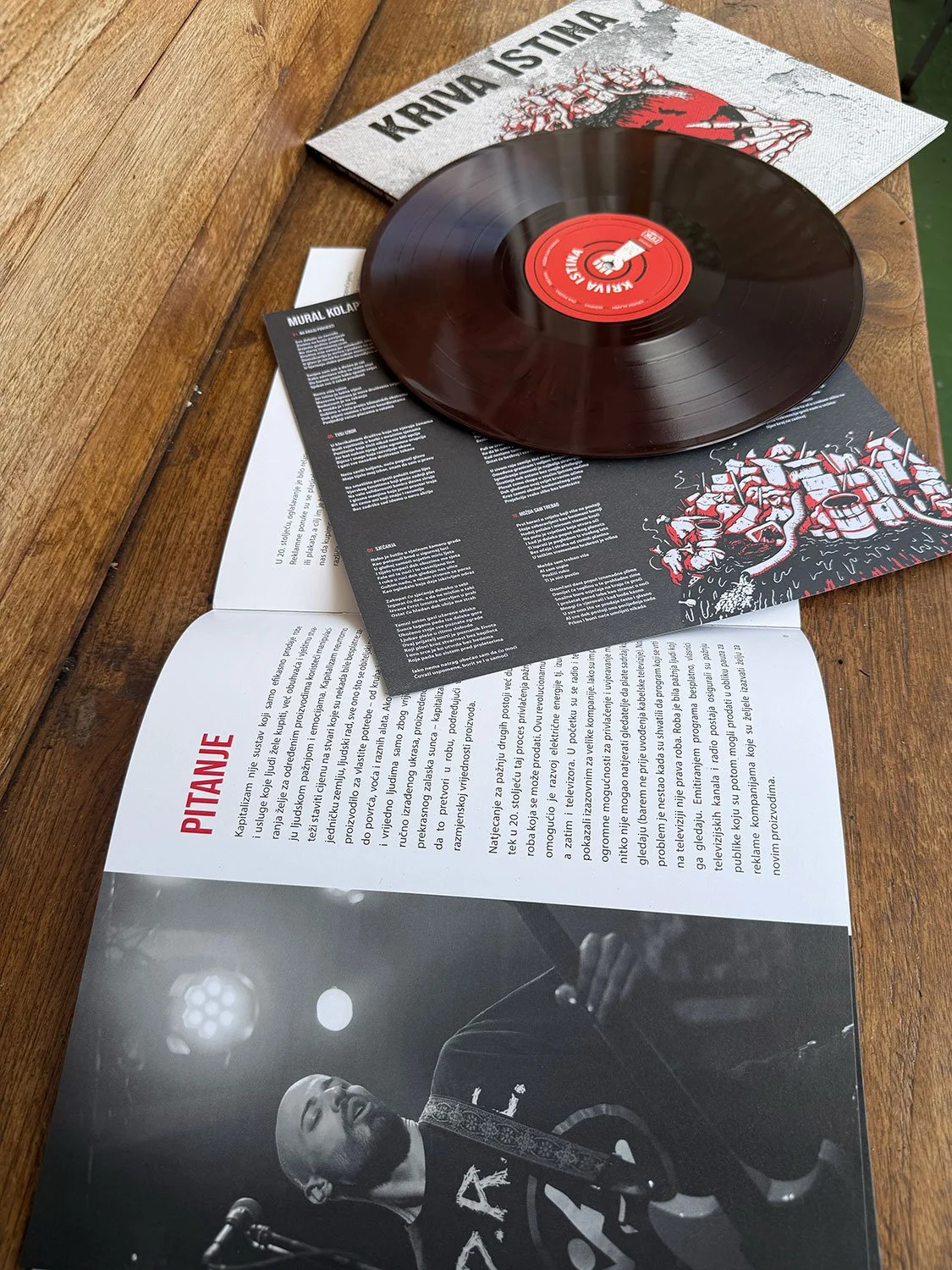 A vinyl record on top of a black and white magazine or booklet with a photo of a rapper holding a microphone. There are also some papers underneath with text, one of which has the word "PITANJE" in red.