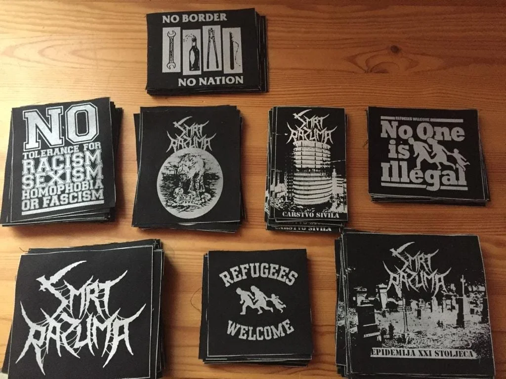 Various black and white political and activist stickers and patches on a wooden surface. Messages include "No Tolerance for Racism, Sexism, Homophobia or Fascism," "No Border, No Nation," "No One is Illegal,""Refugees Welcome," and various artistic d
