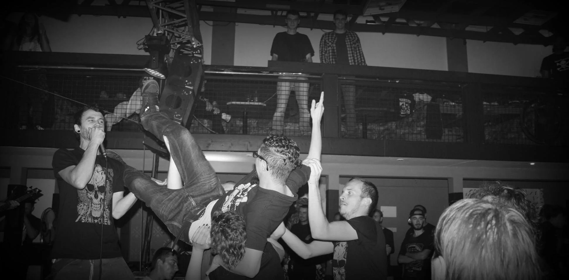 People at a concert scene, with a person being crowd-surfed, holding a guitar, next to a singer with a microphone. The crowd is engaged, with some raising their hands, in an indoor venue with a balcony and visible stage equipment.