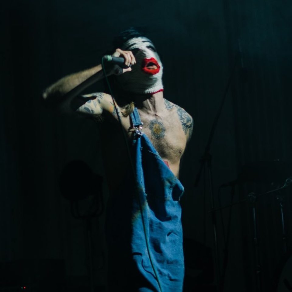A shirtless man with tattoos wearing overalls and clown makeup, holding a microphone and singing on stage in a dark setting.