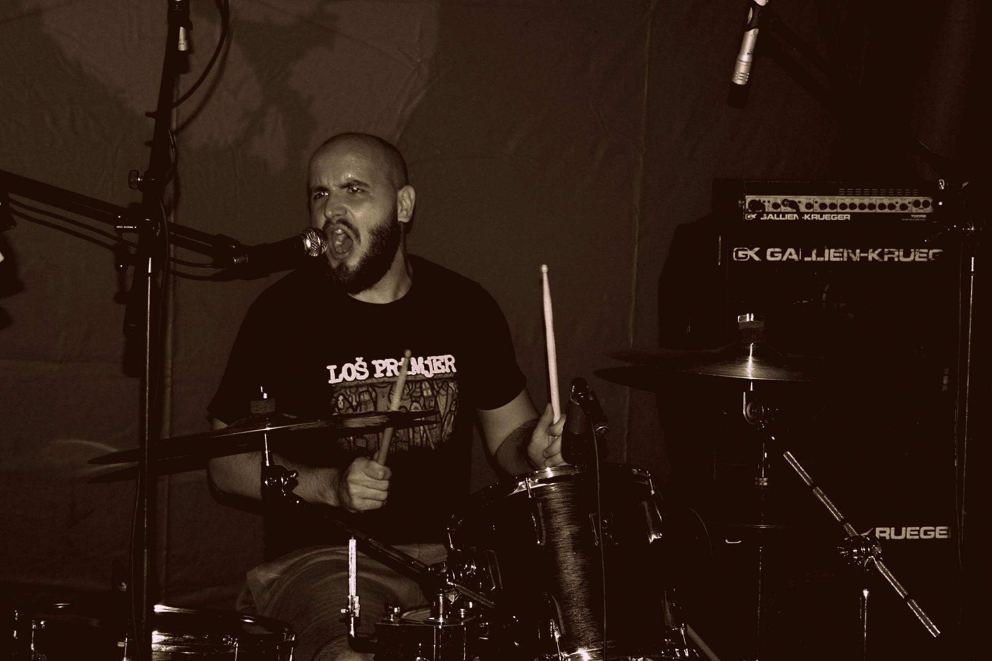 A man with a beard playing drums and singing into a microphone in a dimly lit room, with bass amplifier in the background.