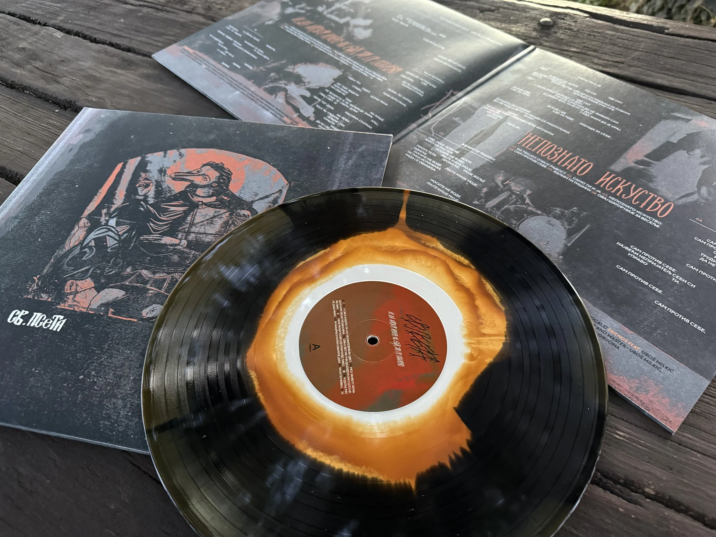 A black vinyl record with a yellow-orange and black label, resting on top of a black and red poster or magazine laid on a wooden surface. The poster features artwork and text in Russian.