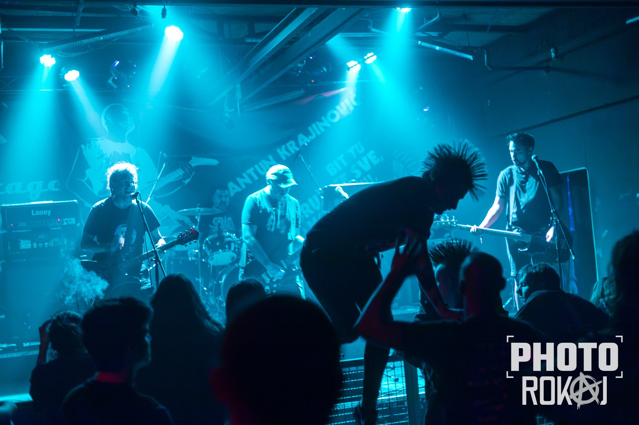 A live band performs on a stage illuminated with blue and purple lights, with a crowd of people dancing in front of them.
