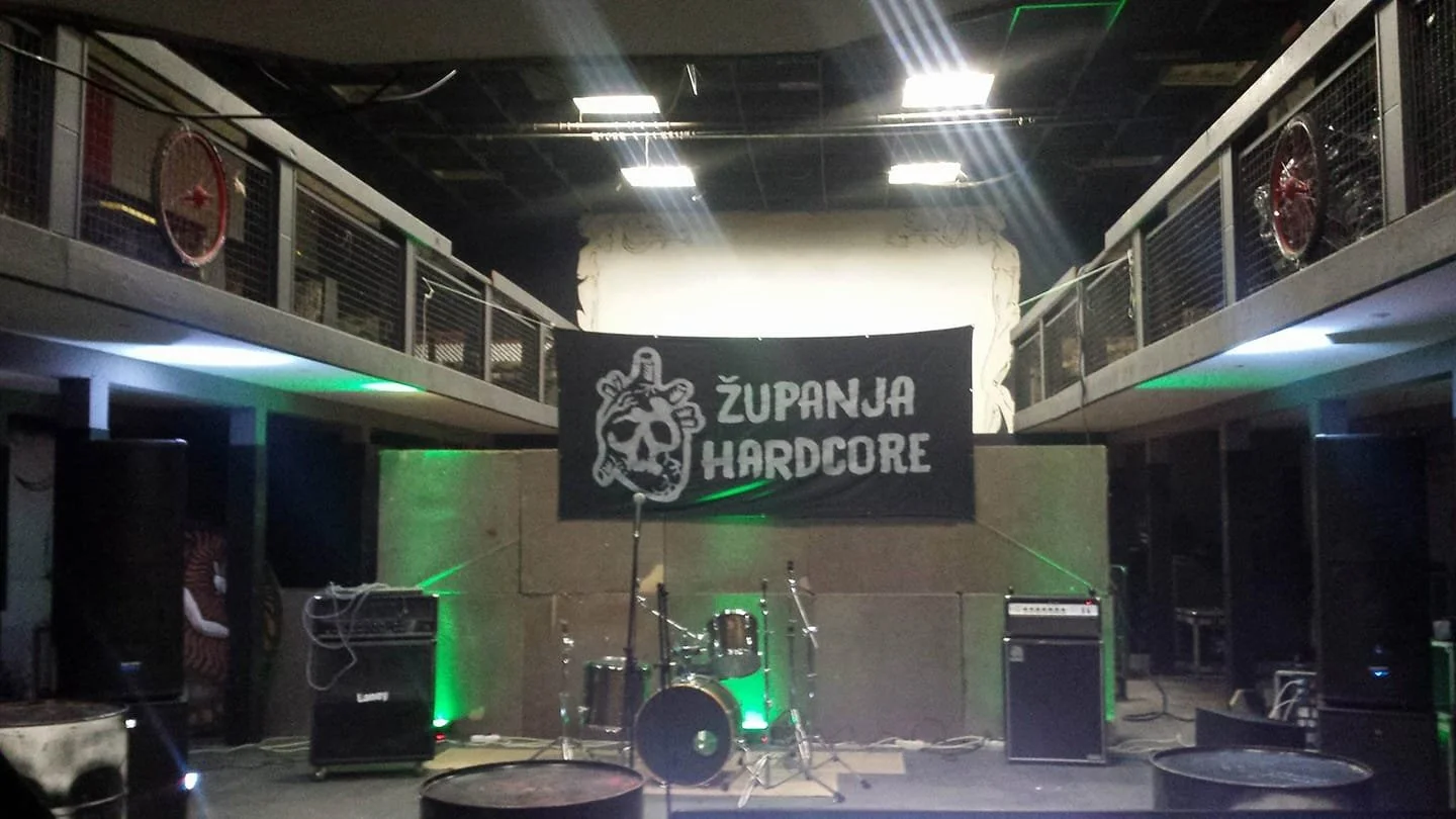 A music stage with drums, amplifiers, speakers, and a microphone, in a dimly lit venue with a banner reading 'ŽUPANJA HARDCORE' hung in the background and green lights illuminating the stage.
