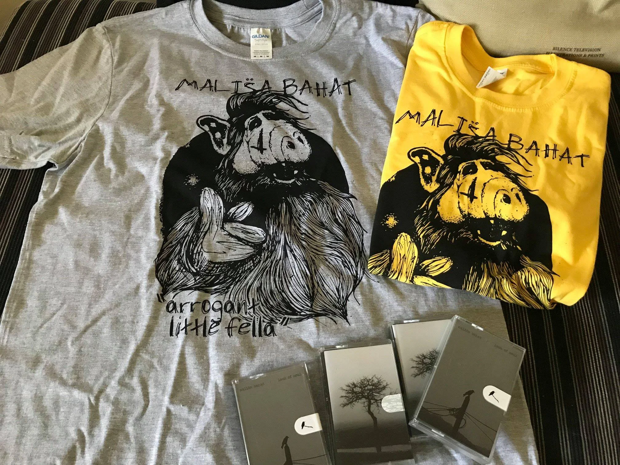 Two T-shirts, one gray and one yellow, both with a black graphic of a lion and the phrase 'Malisa Bahat'. Surrounding the shirts are several small black-and-white photographs of trees and a person holding a leash.