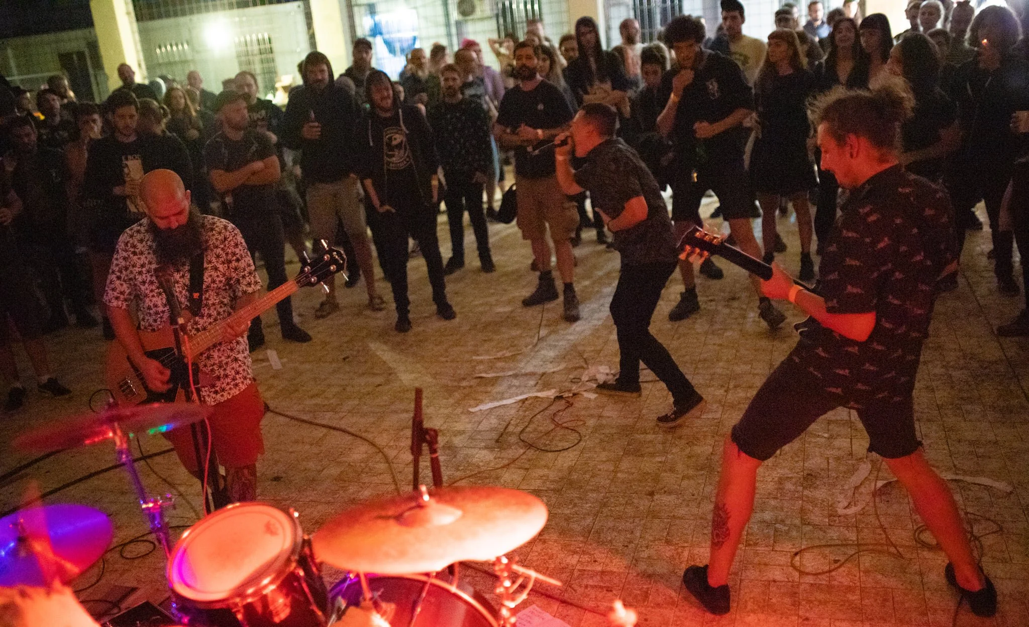 People attending a live music performance in an outdoor or semi-outdoor setting, with musicians playing instruments and a crowd gathered around, some singing and dancing.