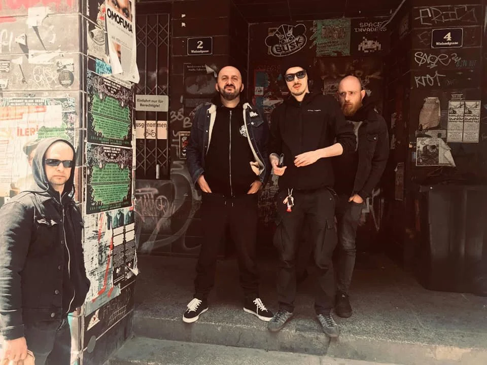 Four men standing in front of a graffiti-covered wall and entrance, wearing dark clothing and sunglasses, with one holding a microphone.
